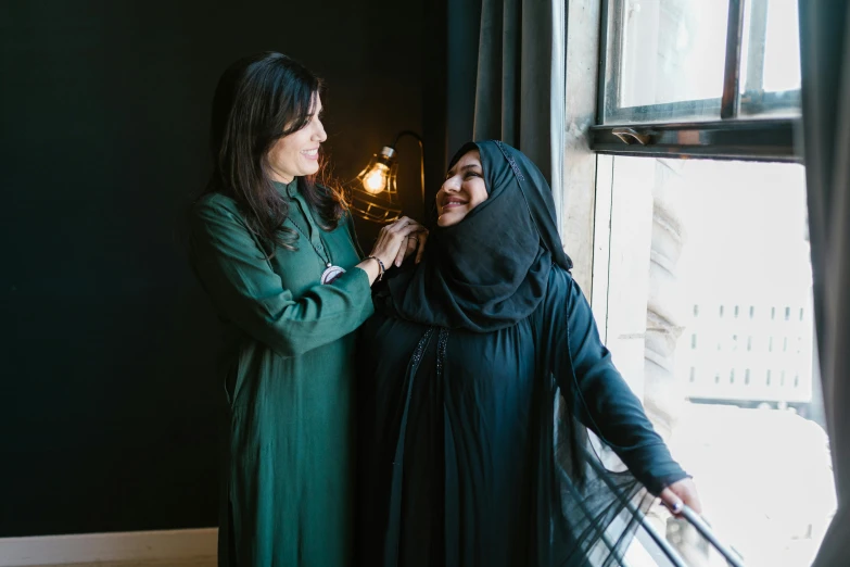 a couple of women standing next to a window, pexels contest winner, hurufiyya, wearing a black robe, smiling at each other, ameera al taweel, green and black