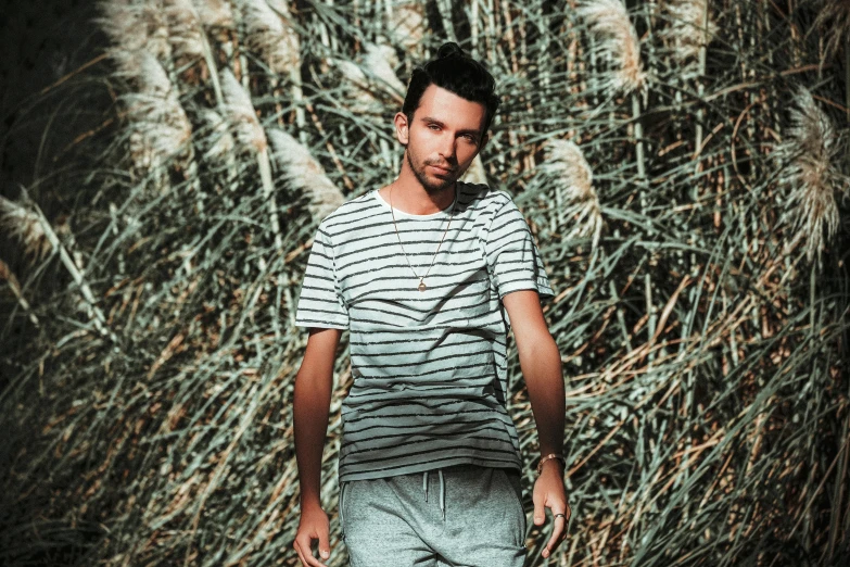 a man standing in front of tall grass, a picture, inspired by Raphaël Collin, unsplash, op art, wearing pants and a t-shirt, young greek man, very handsome, flume