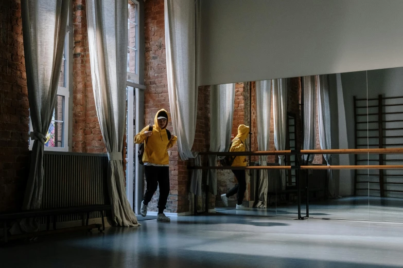 a man in a yellow jacket standing in front of a window, by Nina Hamnett, pexels contest winner, theater dance scene, interior of a loft, wearing a hoodie and sweatpants, standing in class