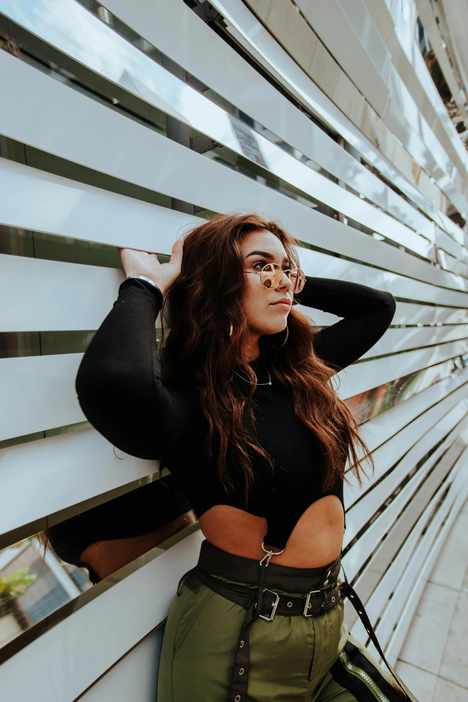 a woman leaning against a wall with her hands on her head, by Robbie Trevino, trending on pexels, wavy long black hair and glasses, wearing a cropped tops, standing astride a gate, wearing black stylish clothing