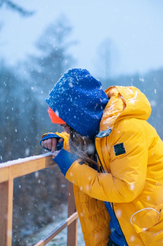 a person standing on a bridge in the snow, in blue and yellow clothes, looking off into the distance, kids, orange reflective puffy coat