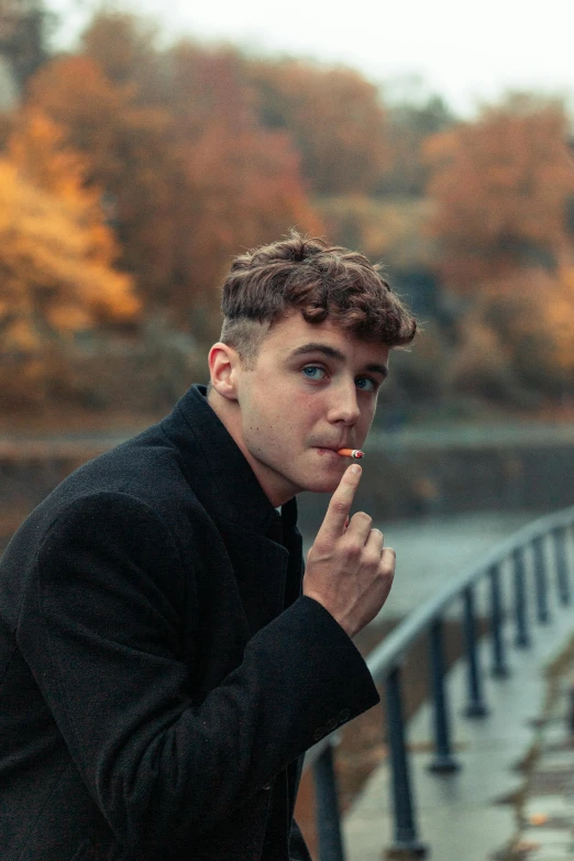 a man standing next to a body of water, an album cover, inspired by Kyle Lambert, pexels contest winner, with a cigarette in its mouth, autumn, cute young man, douglas smith