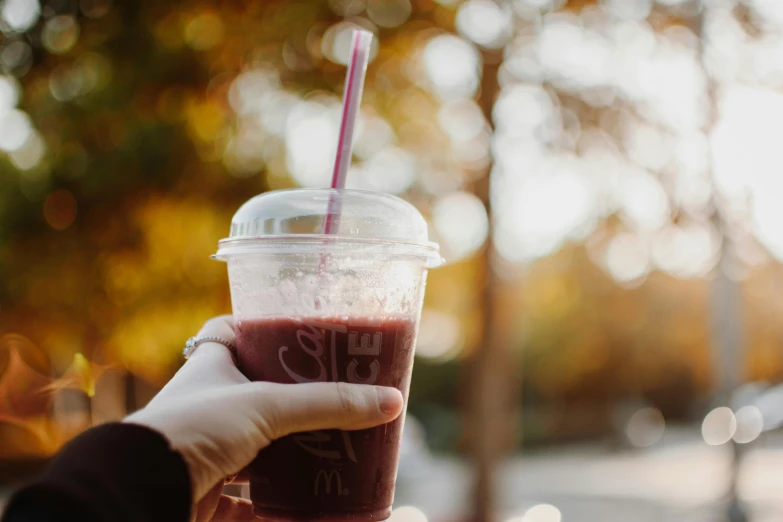 a person holding a plastic cup with a drink in it, by Emma Andijewska, pexels, berry juice, during autumn, milkshake, background image