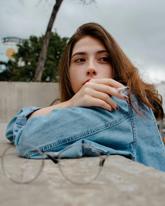 a woman sitting on the ground smoking a cigarette, inspired by Elsa Bleda, trending on pexels, photorealism, denim jacket, wearing square glasses, low quality photo, distant thoughtful look