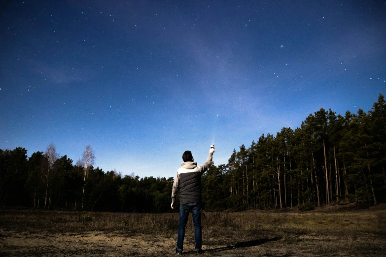 a person standing in a field flying a kite, by Julia Pishtar, pexels contest winner, forest at night, holding a blue lightsaber, men look up at the sky, pointing