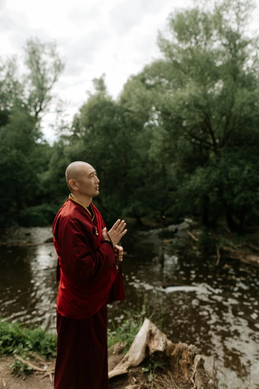 a man in a red robe standing in front of a river, meditating, against the backdrop of trees, ukrainian monk, darren quach