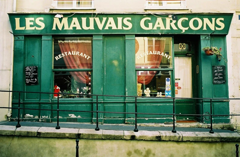 a green building with a sign that says les mauvais garcons, an album cover, inspired by Maurice Utrillo, pexels contest winner, food stall, gray, exterior, decadent