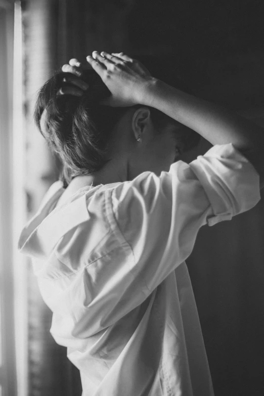 a woman standing in front of a window with her hands on her head, a black and white photo, unsplash, clothed in white shirt, struggling, photo of the girl, hair tied back