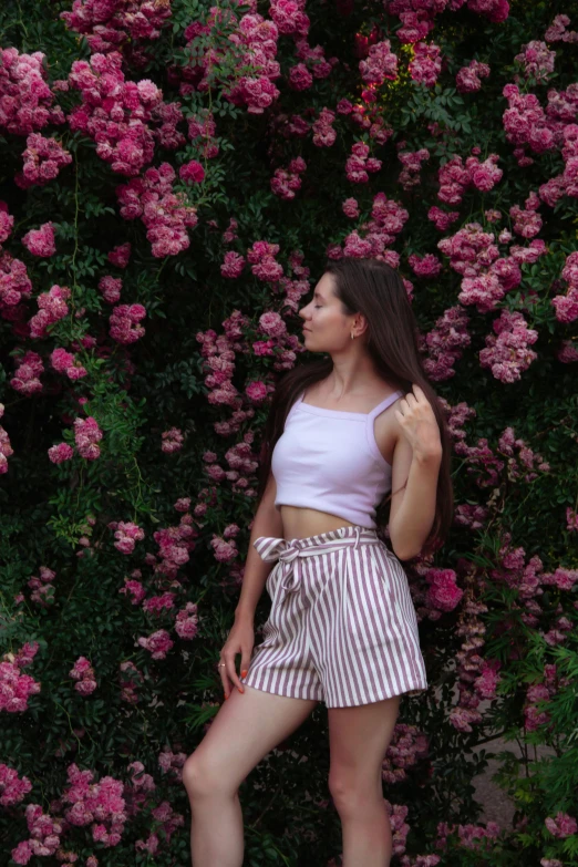 a woman standing in front of a bush of pink flowers, pexels contest winner, croptop and shorts, stripes, anna nikonova aka newmilky, 18 years old
