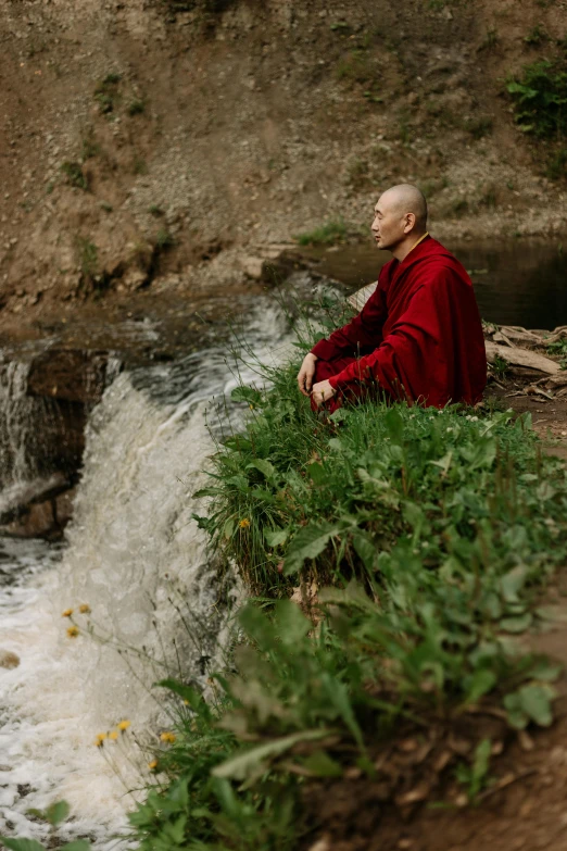 a man sitting on top of a lush green hillside next to a river, a portrait, inspired by Steve McCurry, unsplash, renaissance, wearing red robes, buddhist monk meditating, portrait tilda swinton, draped with water and spines