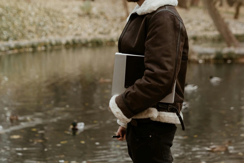 a man standing in front of a body of water, an album cover, trending on pexels, with a laptop on his lap, leather fur jacket, in a park, holding notebook