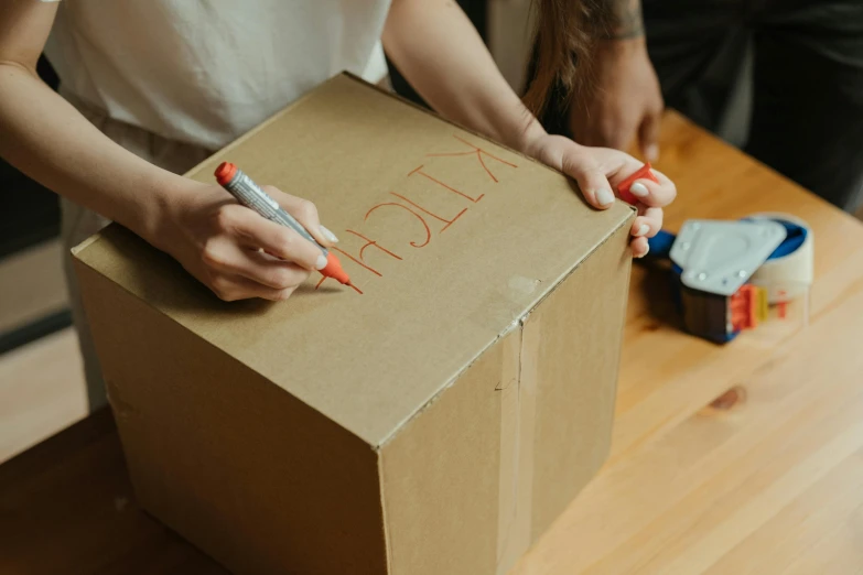 a woman is writing on a cardboard box, pexels contest winner, 🦩🪐🐞👩🏻🦳, permanent marker, furniture, brown