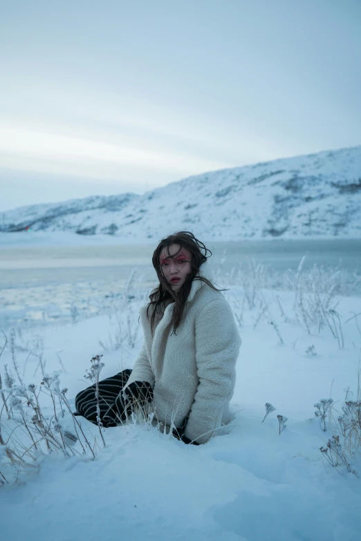 a woman sitting in the snow next to a body of water, an album cover, inspired by Elsa Bleda, trending on pexels, land art, woman in a sheep costume, actress, cold blood, nordic