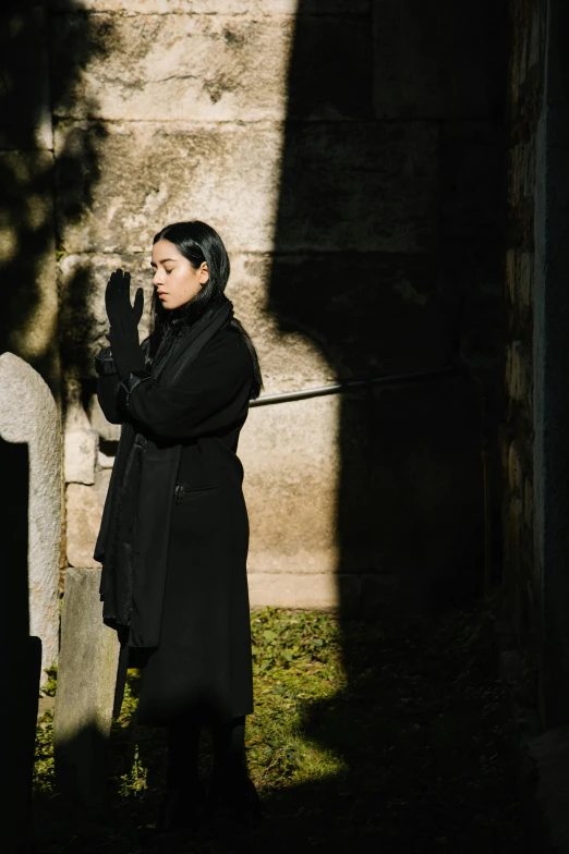 a woman in a black coat standing next to a grave, inspired by Modest Urgell, pexels contest winner, symbolism, gong li, hood and shadows covering face, ignant, ( ( theatrical ) )