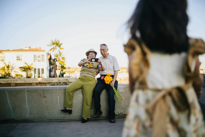 a man and a woman sitting next to each other, by Nathalie Rattner, pexels contest winner, happening, wearing yellow floral blouse, people watching around, carrying flowers, an oldman