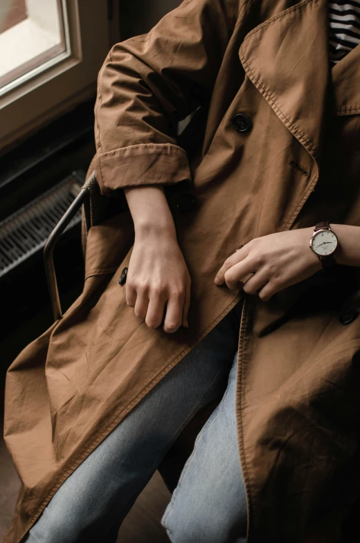 a woman sitting on a bench next to a window, trending on pexels, light brown trenchcoat, wears a watch, roots and hay coat, closeup of hand