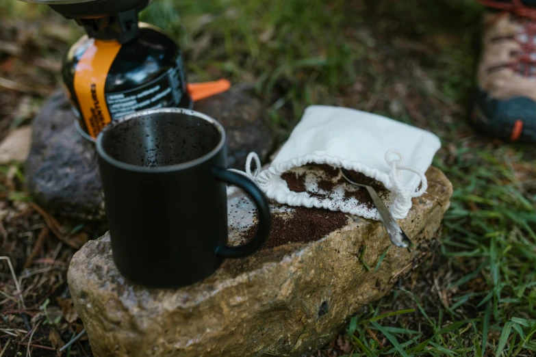 a cup of coffee sitting on top of a rock, pouches, with a white mug, wilderness ground, detailed product image