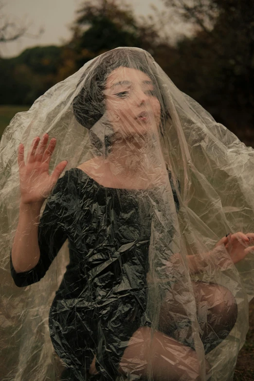 a woman standing in a field covered in plastic, inspired by Marina Abramović, unsplash, performance, bubble goth, pudica gesture bouguereau style, while it's raining