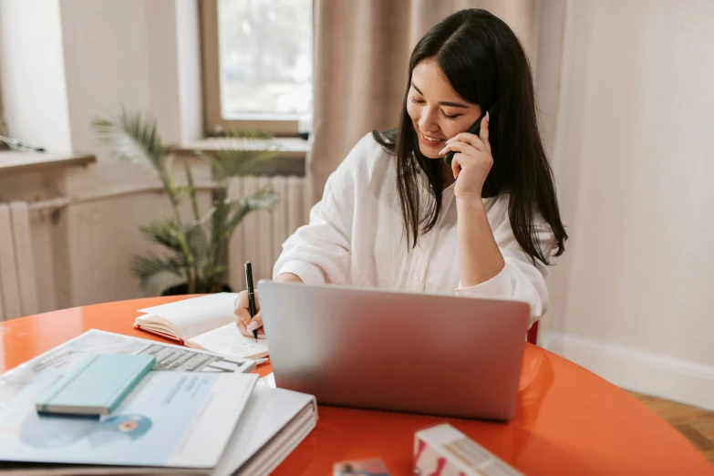 a woman sitting at a table talking on a cell phone, working on her laptop, selling insurance, brown and white color scheme, an asian woman