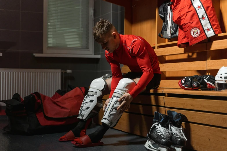 a man sitting on a bench in a locker, inspired by Jakub Husnik, shutterstock, full ice hockey goalie gear, red boots, instagram picture, wearing knee and elbow pads