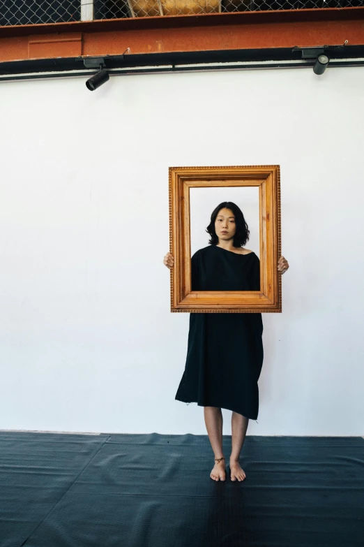 a woman holding a picture frame in front of her face, inspired by Marina Abramović, unsplash, visual art, full - length portrait, asian women, square, wearing a dark dress