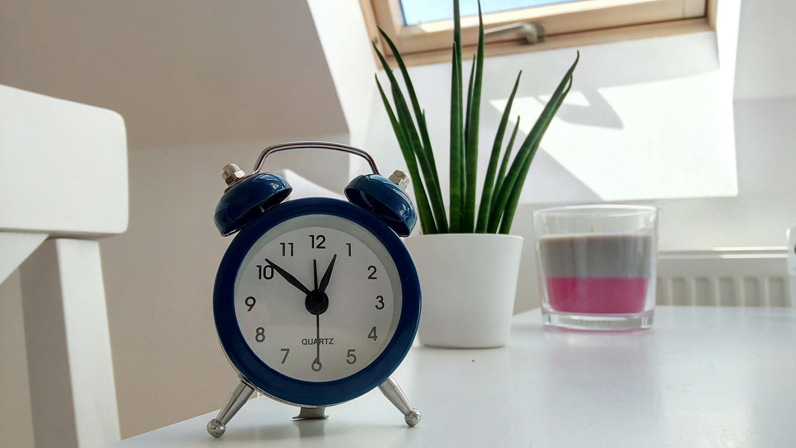 a blue alarm clock sitting on top of a white table, next to a plant, small and cosy student bedroom, blue and grey, clocks