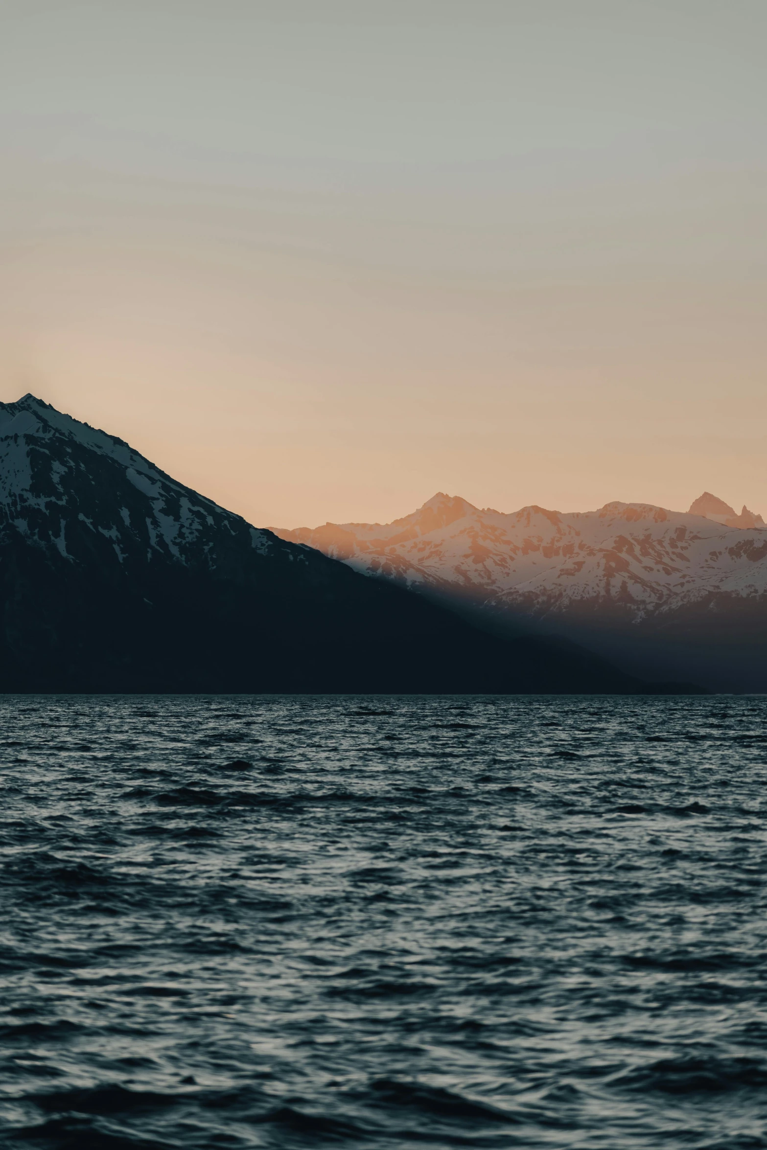 a large body of water with mountains in the background, by Johannes Voss, unsplash contest winner, cold sunset, viewed from the ocean, low quality photo, snow capped mountains