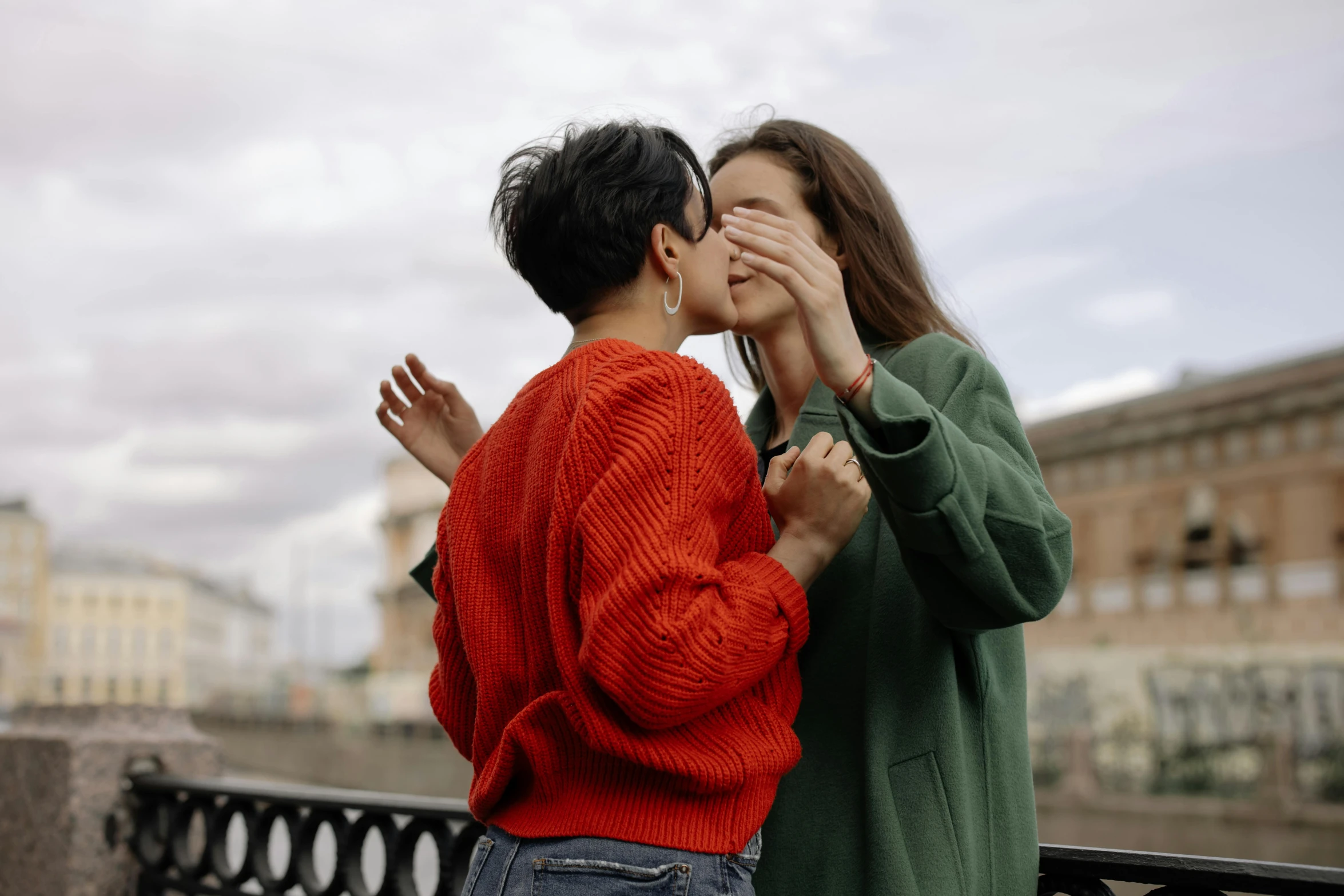 a couple standing next to each other on a bridge, trending on pexels, antipodeans, lesbian kiss, wearing a red turtleneck sweater, hands shielding face, celebrating