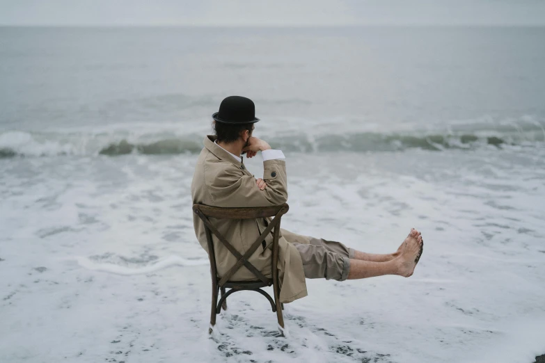 a man sitting in a chair on the beach, a portrait, inspired by Hugo Simberg, unsplash, on a rainy day, ignant, colin hay, invisible man