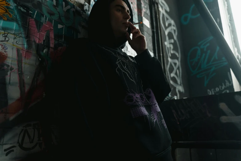 a woman talking on a cell phone in front of a graffiti covered wall, a picture, by Daarken, unsplash, graffiti, young man in a purple hoodie, ominous gothic aesthetic, black hoodie, **cinematic