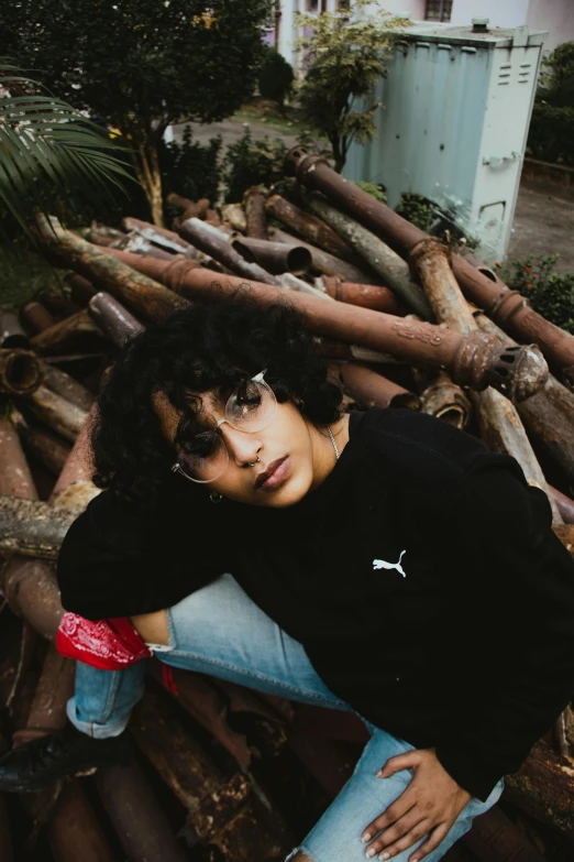 a woman sitting on top of a pile of wood, an album cover, trending on pexels, (dark shorter curly hair), androgynous male, assamese aesthetic, taken with sony alpha 9