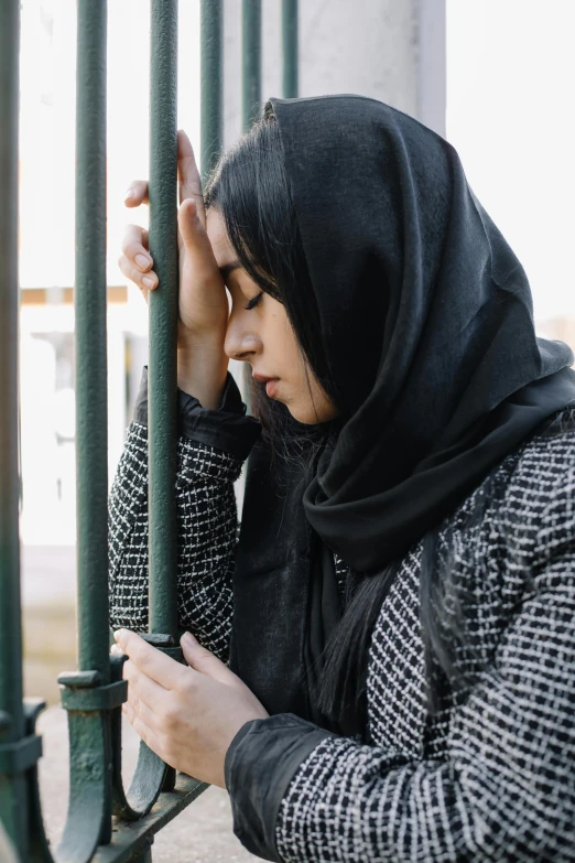 a woman leaning against a fence looking at her cell phone, by Maryam Hashemi, trending on pexels, hurufiyya, wrapped in a black scarf, distraught, standing astride a gate, head bowed slightly