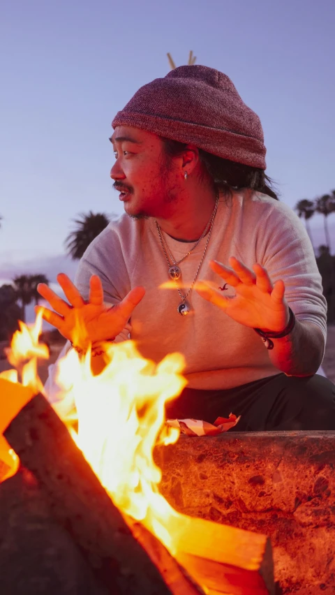 a man sitting next to a fire with palm trees in the background, an album cover, inspired by Kanō Tan'yū, unsplash, talking around a fire, david choe, press photos, joe taslim