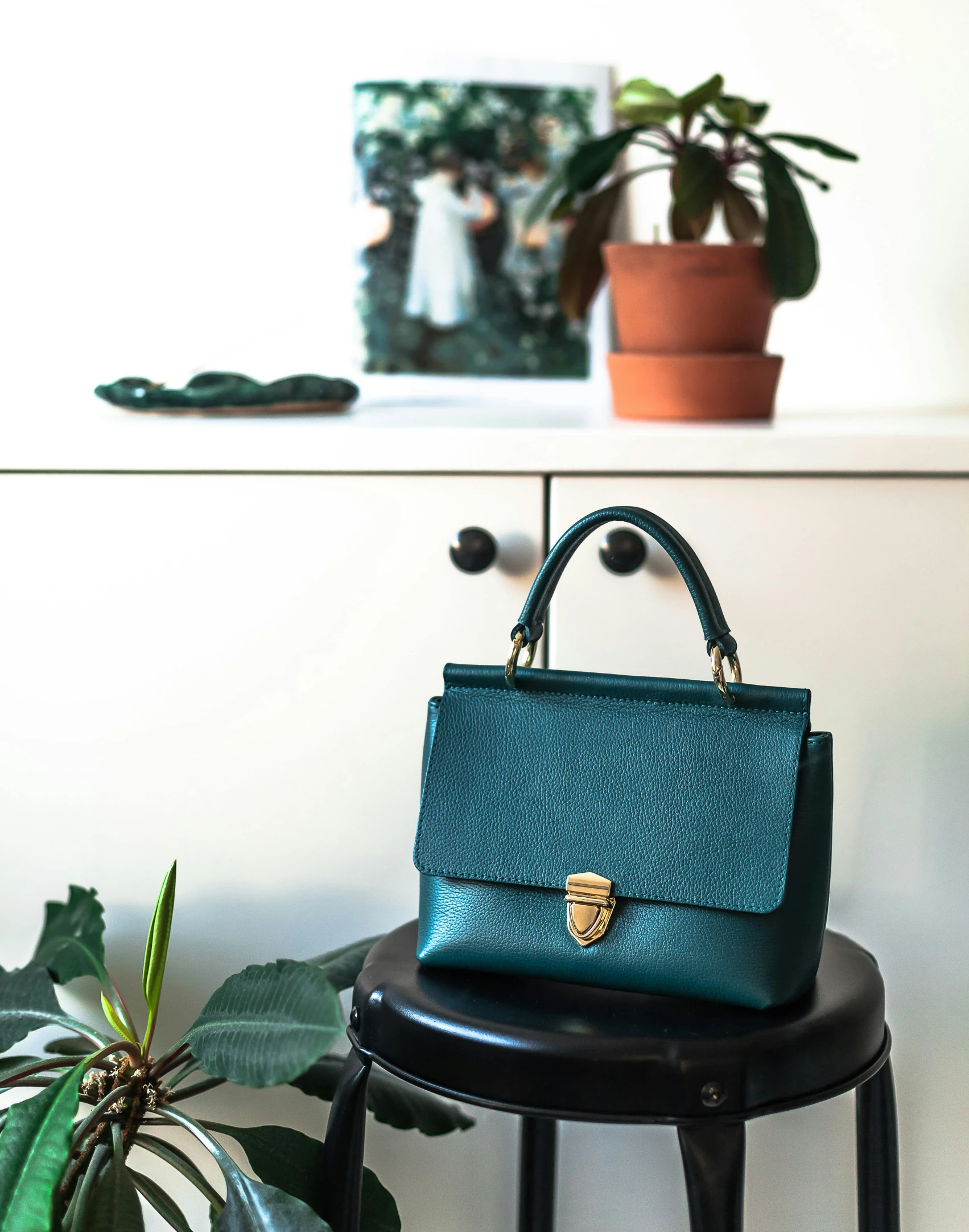 a purse sitting on top of a stool next to a potted plant, by Julia Pishtar, featured on instagram, in a dark teal polo shirt, official product photo, elegant lady, fully frontal view