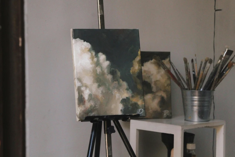 a painting sitting on top of a wooden easel, inspired by Thomas Wijck, unsplash, tonalism, volumetric clouds, some square paintings, in style of wlop, on canvas