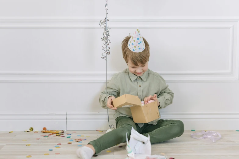 a little boy sitting on the floor holding a box, by Eden Box, happening, wearing a party hat, grey, multi - coloured, plain