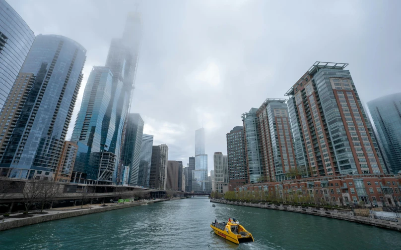 a yellow boat traveling down a river next to tall buildings, a picture, by Robbie Trevino, chicago, 2022 photograph, foggy!, tubing