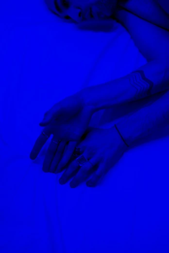 a woman laying on top of a bed under a blue light, an album cover, inspired by Yves Klein, unsplash, symbolism, corrected hands, blacklight aesthetic, laying down with wrists together, low fi