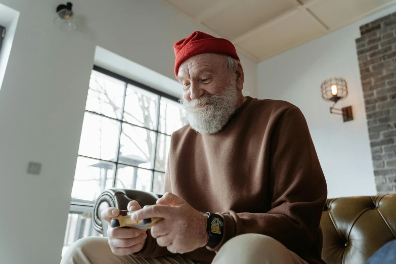 a man sitting on a couch looking at his cell phone, by Jesper Knudsen, pexels contest winner, hyperrealism, red cap, white beard, brown, holding controller