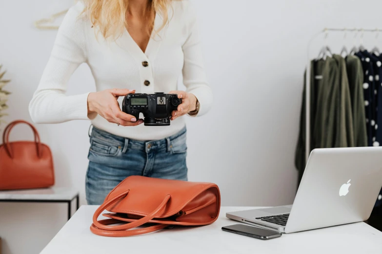 a woman holding a camera in front of a laptop, holding a leather purse, photoshoot for skincare brand, on a white table, middle shot waist up