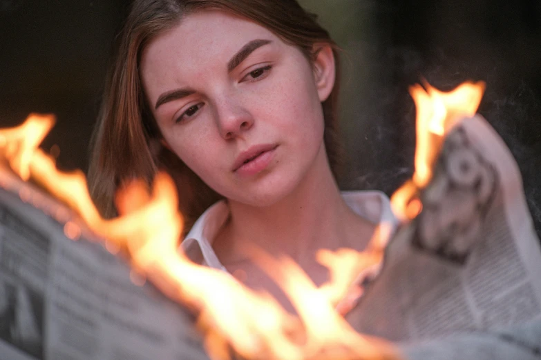 a woman reading a newspaper with flames coming out of it, by Julian Allen, pexels contest winner, realism, portrait of anya taylor-joy, outdoor campfire pit, close up face, teenager girl