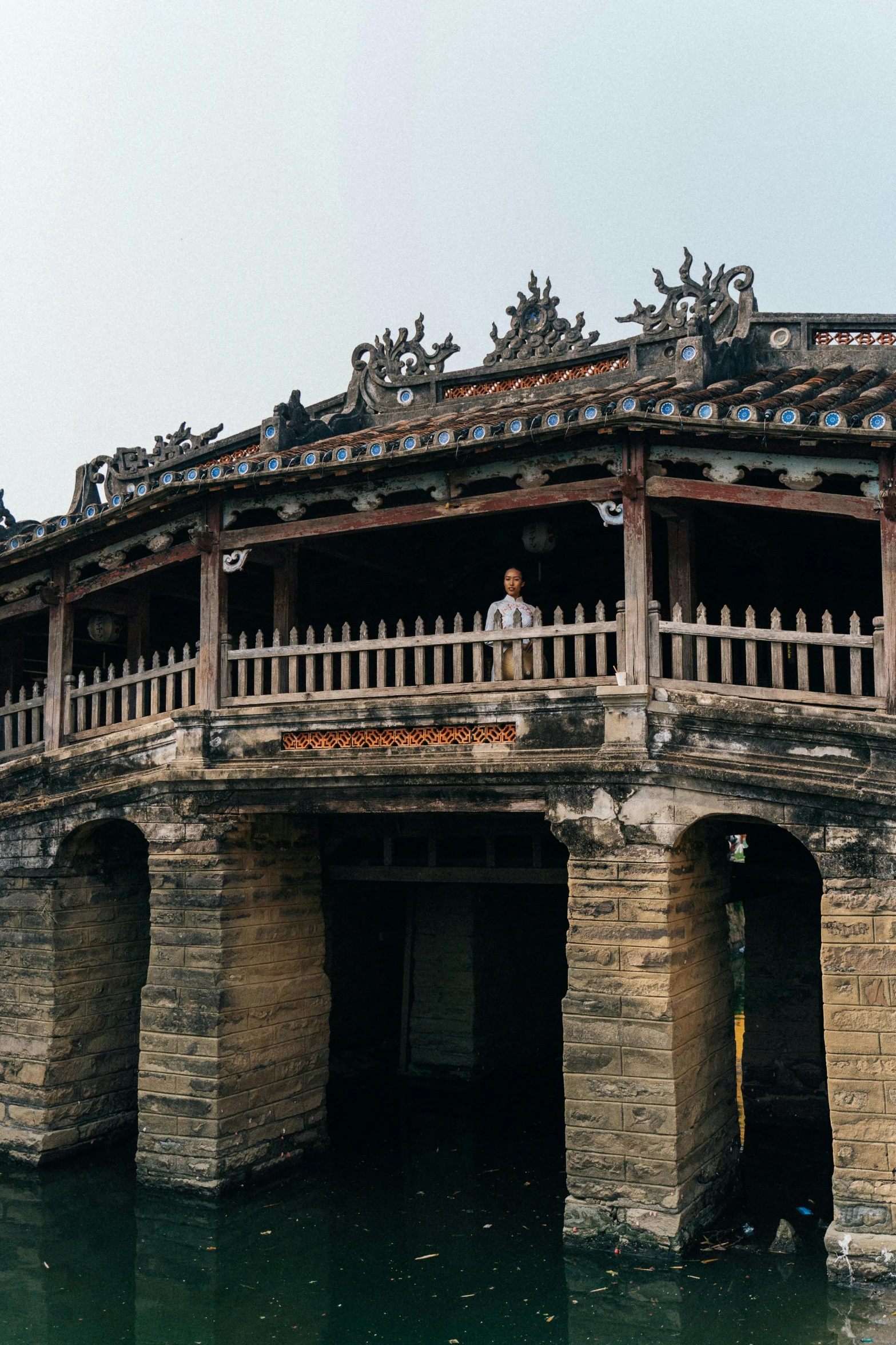 a stone bridge over a body of water, inspired by Gu An, elaborate carved wood balconies, vietnamese woman, panoramic, rounded roof