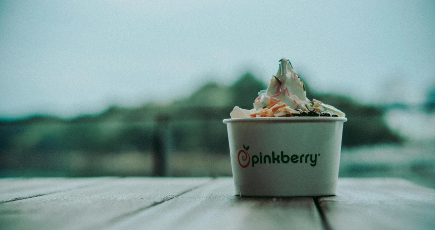 a cup of ice cream sitting on top of a wooden table, inspired by Per Kirkeby, graffiti, the sky is pink, its name is greeny, berries inside structure, promo image