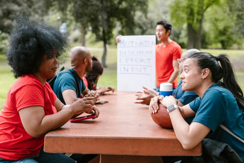 a group of people sitting around a wooden table, by Julia Pishtar, at a park, interrupting the big game, woman holding sign, varying ethnicities