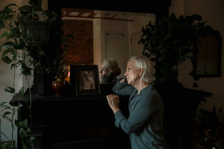 a woman standing in front of a mirror in a living room, a portrait, pexels contest winner, photorealism, dark grey haired man, praying, in front of a fireplace, white haired