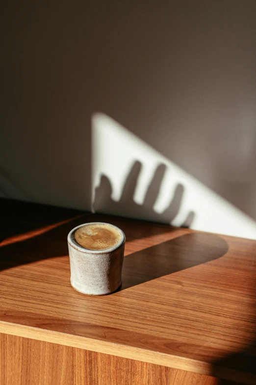 a cup of coffee sitting on top of a wooden table, by Rebecca Horn, long cast shadows, ignant, detailed product image, suns