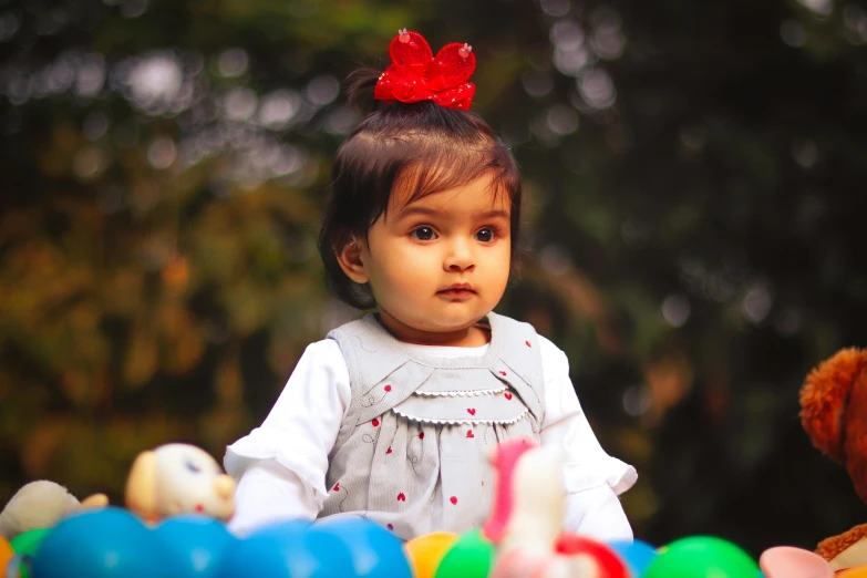 a little girl sitting on top of a pile of toys, by irakli nadar, pexels contest winner, serious focussed look, balloon, at the park, a red bow in her hair