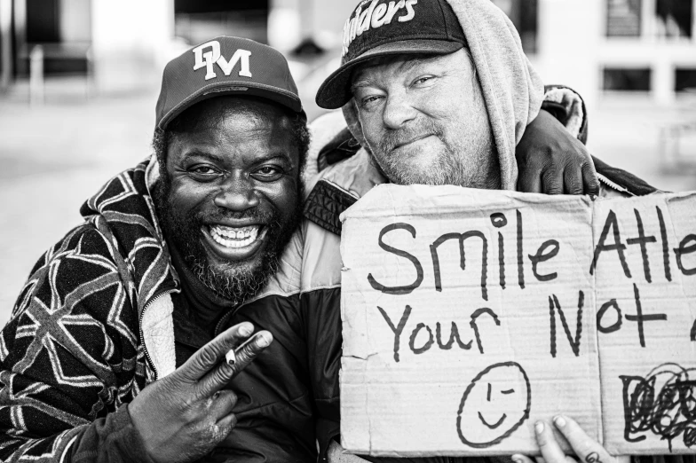 a couple of men sitting next to each other holding a sign, a black and white photo, by Matija Jama, pexels, detailed realistic smiling faces, skilled homeless, smiley face, portrait of mf doom