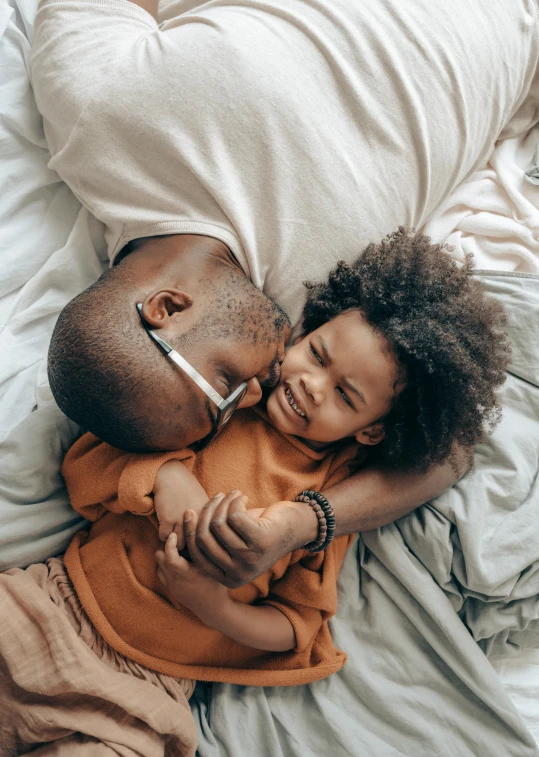 a man laying on top of a bed next to a child, a stock photo, pexels contest winner, symbolism, african american girl, holding each other, arm around her neck, hugging each other