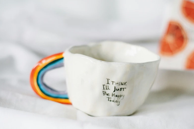 a coffee cup sitting on top of a white table, a child's drawing, inspired by Tracey Emin, featured on instagram, happening, bright rainbow nimbus, hand built ceramics, on grey background, dua lipa